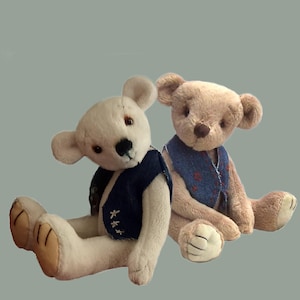 Peut inclure: Deux ours en peluche assis côte à côte. L'ours de gauche est de couleur crème avec un gilet bleu foncé orné de détails cousus blancs. L'ours de droite est brun clair et porte un gilet bleu à motifs avec de petits accents floraux rouges.