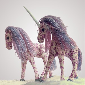 Può includere: Due giocattoli di peluche a forma di unicorno con tessuto floreale rosa e bianco e capelli in lana viola e bianco. L'unicorno più grande ha un corno bianco.
