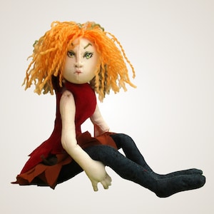 Könnte beinhalten: Eine handgemachte Puppe mit orangefarbenen Locken, einem roten Kleid und einer blauen Hose. Die Puppe sitzt mit gekreuzten Beinen und die Arme liegen auf ihrem Schoß.
