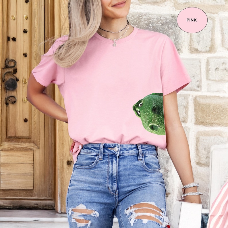 May include: A light pink short-sleeved t-shirt featuring a graphic of a green dinosaur head peeking from the lower right side. The shirt has a crew neckline and a solid color design. A small circular pink tag with the word PINK is visible.