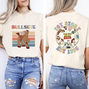 Może przedstawiać: Kremowa koszulka z grafiką Bullseye i napisem "BULLSEYE". Tył koszulki zdobi okrągły wz&oacute;r z napisem "TOY STORY EST 1995" i postaciami z Toy Story.