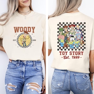 K&ouml;nnte beinhalten: Cremefarbenes T-Shirt mit Woody aus Toy Story und dem Text "WOODY EST. 1999" auf der Vorderseite. Die R&uuml;ckseite des Shirts zeigt eine Gruppe von Toy Story-Charakteren auf kariertem Hintergrund mit dem Text "TOY STORY - Est. 1999".