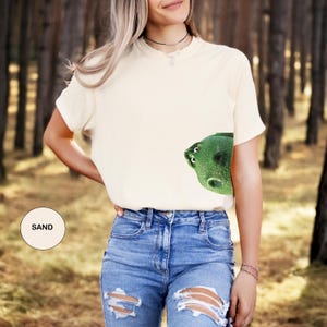 May include: A sand-colored short-sleeve t-shirt featuring a graphic of a green, textured, wide-eyed creature peeking from the lower right side. The shirt has a crew neckline and a plain back. A small circular label indicates the color is sand.