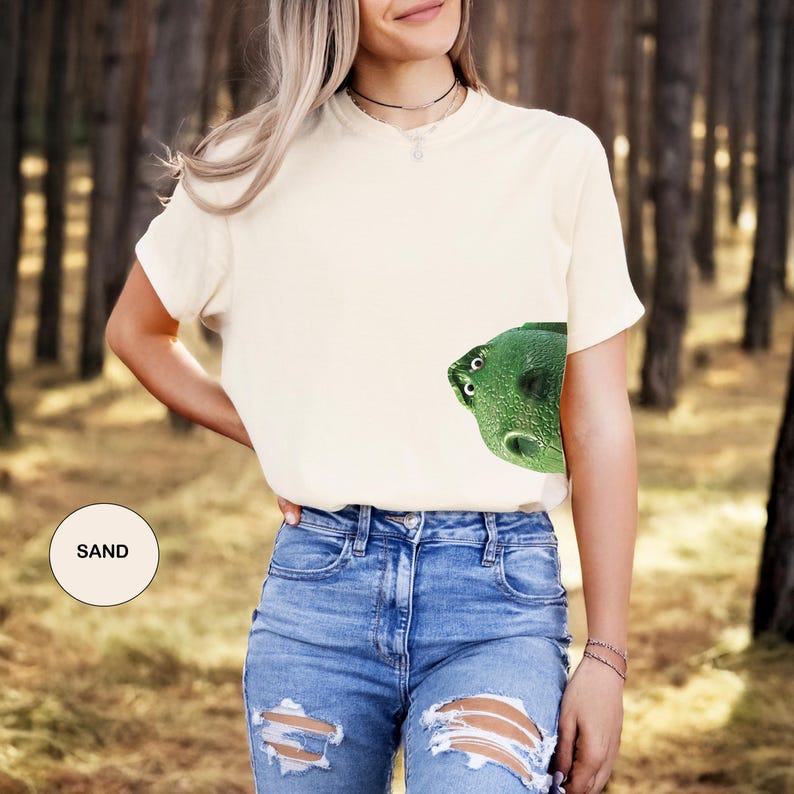 May include: A sand-colored short-sleeve t-shirt featuring a graphic of a green, textured, wide-eyed creature peeking from the lower right side. The shirt has a crew neckline and a plain back. A small circular label indicates the color is sand.
