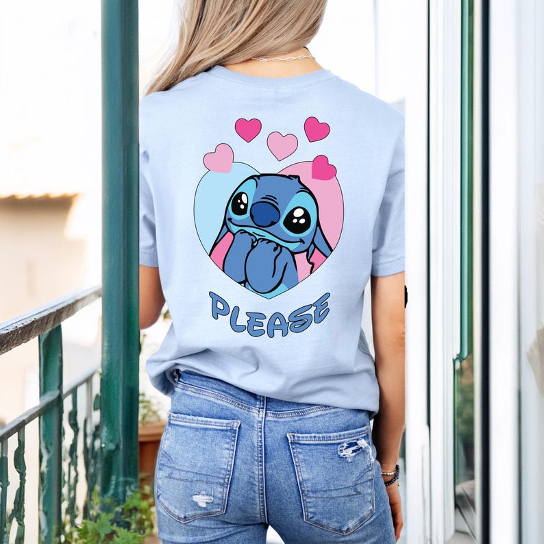 May include: Light blue t-shirt featuring a cartoon character inside a heart-shaped design with pink hearts above. The word "PLEASE" is printed below the character. The shirt is made of a soft material.