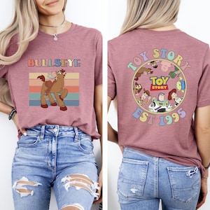Może przedstawiać: Malinowa koszulka z grafiką Bullseye i napisem "BULLSEYE". Tył koszulki zdobi wz&oacute;r "Toy Story" z postaciami i napisem "EST 1995". Zabawny, kolorowy design dla fan&oacute;w filmu.