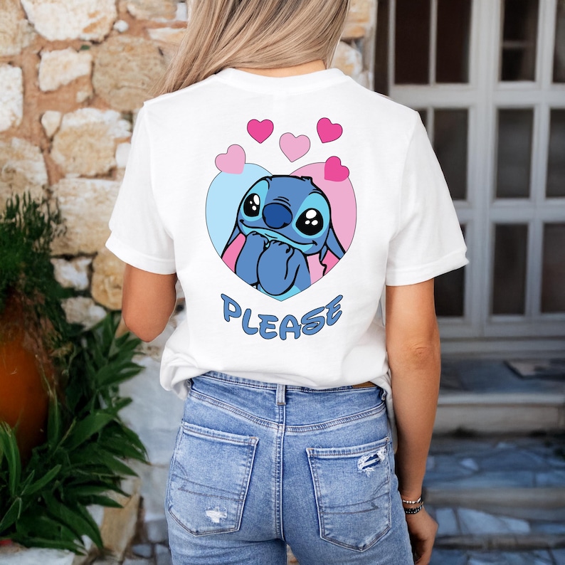 May include: White t-shirt featuring a blue cartoon character inside a heart-shaped design with pink and blue accents. The word "PLEASE" is written in blue below the heart. The shirt is paired with blue jeans.