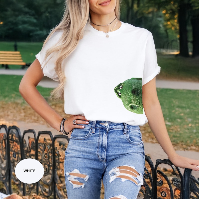 May include: A white short-sleeved t-shirt featuring a graphic print of a green, textured creature with large white eyes peeking out from the lower right side. The shirt is paired with blue denim jeans that have distressed, ripped detailing on the thighs.