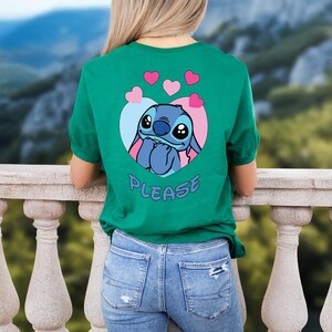 May include: Green t-shirt featuring a cartoon character inside a heart-shaped design with pink and blue accents. The word "PLEASE" is printed below the character. The shirt is made of a soft material and is suitable for casual wear.