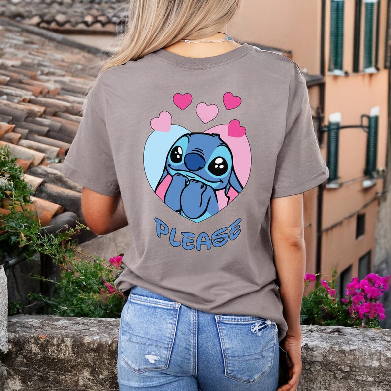 May include: A gray t-shirt featuring a cartoon character within a heart-shaped design. The character is blue with large eyes and is surrounded by pink and blue accents. The word "PLEASE" is written in blue below the character.