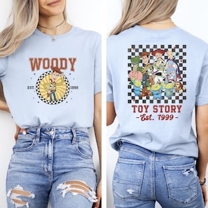 K&ouml;nnte beinhalten: Hellblaues T-Shirt mit Woody von Toy Story, dem Text "WOODY" und "EST. 1999" auf der Vorderseite. Die R&uuml;ckseite des Shirts zeigt eine Toy Story-Charaktergrafik mit dem Text "TOY STORY -Est. 1999-".