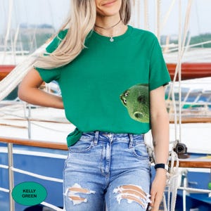 May include: A kelly green short-sleeve t-shirt featuring a graphic of a green frog's head peeking out from the lower right side. The shirt has a crew neckline and is paired with blue denim jeans that have distressed rips at the knees.