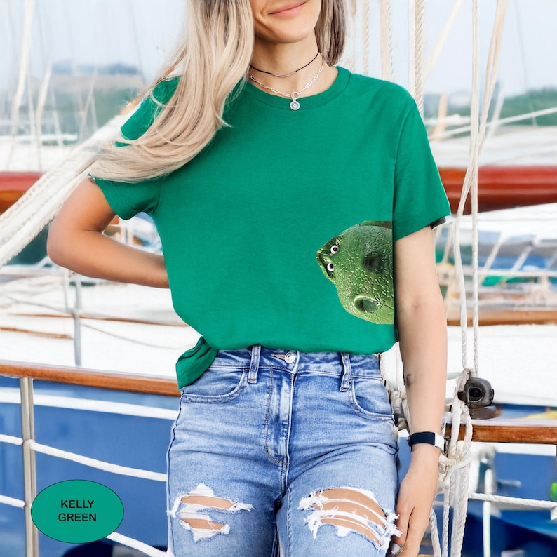 May include: A kelly green short-sleeve t-shirt featuring a graphic of a green frog's head peeking out from the lower right side. The shirt has a crew neckline and is paired with blue denim jeans that have distressed rips at the knees.