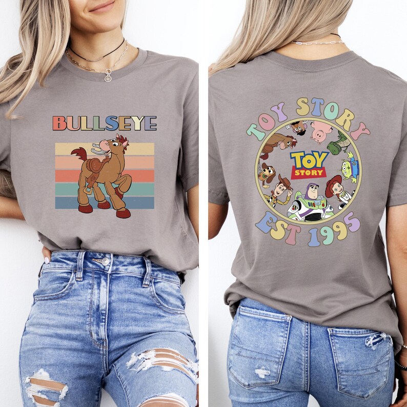 Może przedstawiać: Szara koszulka z grafiką Bullseye'a, konia, z retro wzorem zachodu słońca. Tył koszulki przedstawia okrągły wz&oacute;r Toy Story z postaciami i napisem "EST 1995".