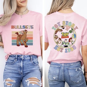 Może przedstawiać: Jasnor&oacute;żowa koszulka z kresk&oacute;wkowym wizerunkiem konia Bullseye i napisem "BULLSEYE". Tył koszulki ma okrągły wz&oacute;r z napisem "TOY STORY" i rokiem "EST 1995".