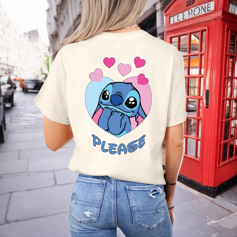 May include: Cream-colored t-shirt featuring a blue cartoon character inside a heart-shaped design with pink and blue accents. The word "PLEASE" is printed below the character. The shirt is paired with blue jeans.