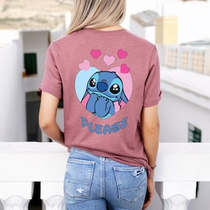 May include: A mauve t-shirt featuring a cartoon character inside a heart-shaped design. The character is blue with large eyes and is surrounded by pink and blue accents. The word "PLEASE" is written in blue below the character.