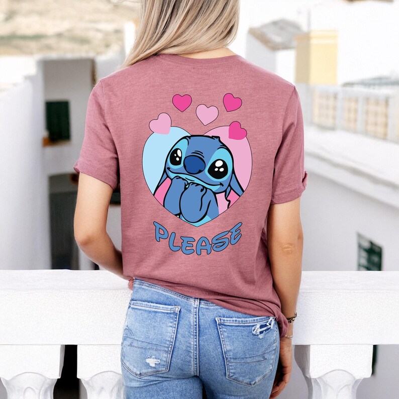 May include: A mauve t-shirt featuring a cartoon character inside a heart-shaped design. The character is blue with large eyes and is surrounded by pink and blue accents. The word "PLEASE" is written in blue below the character.