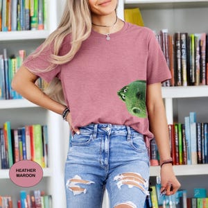 May include: A heather maroon short-sleeve t-shirt featuring a graphic of a green dinosaur head peeking out from the side. The shirt has a crew neckline and is paired with light blue denim jeans that have distressed rips at the knees.