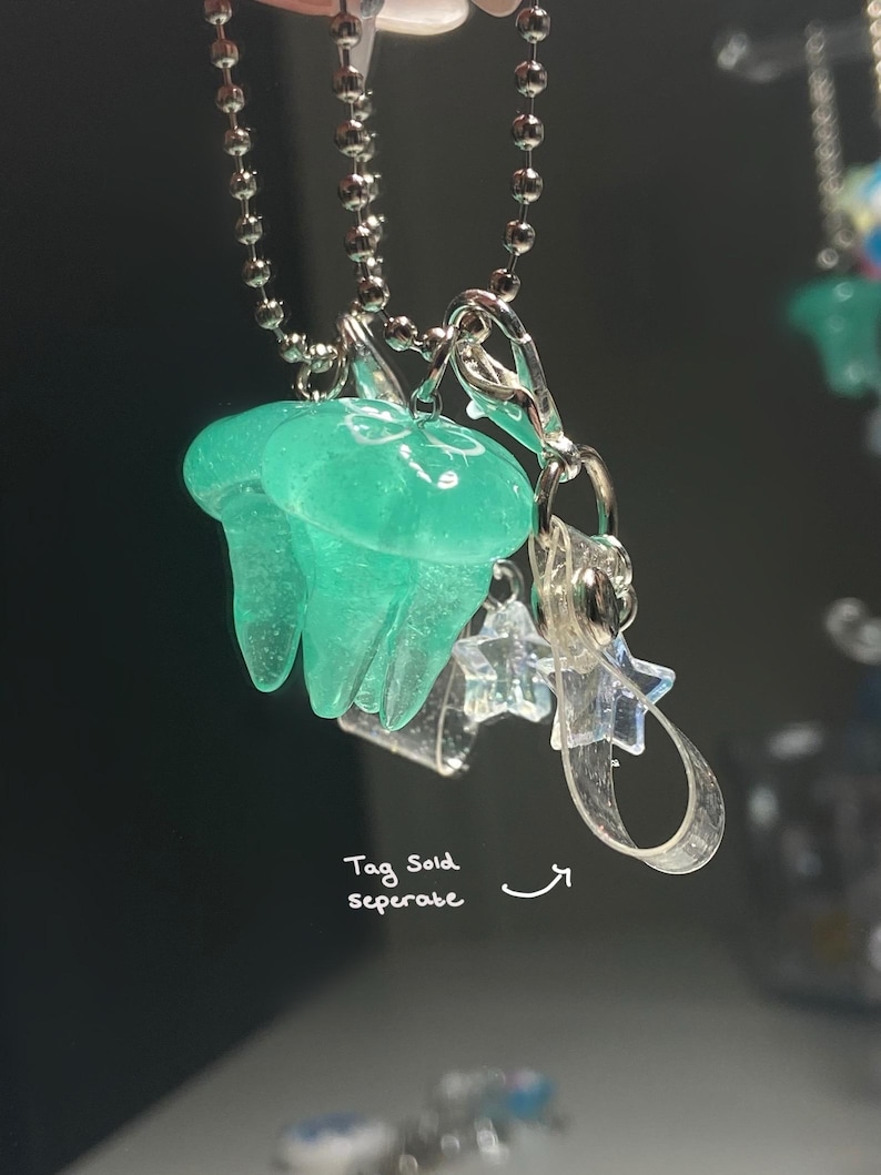 May include: A pair of translucent teal jellyfish charms hanging from a silver ball chain. Clear star-shaped charms and a silver clasp are also attached. The text "Tag Sold separate" is visible.
