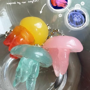 May include: Colorful jellyfish-shaped keychains in yellow, orange, pink, and teal. The keychains are inspired by real jellyfish, as indicated by the text in the image. The keychains are attached to silver chains.