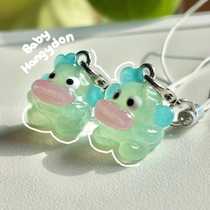 May include: Two translucent mint green charms featuring the character Hangyodon with a pink mouth, black eyes, and blue accents. Each charm has a silver-tone metal lobster clasp attached to a white cord. The text Baby Hangyodon is visible.