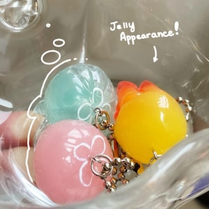 May include: A clear plastic container holds three colorful, round charms with silver-colored chains. The charms are light blue, pink, and yellow, each with a white floral design. The text "Jelly Appearance!" is written above the charms.