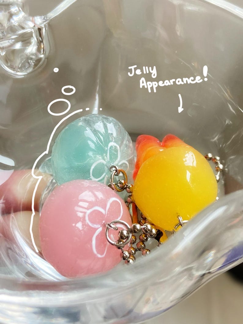 May include: A clear plastic container holds three colorful, round charms with silver-colored chains. The charms are light blue, pink, and yellow, each with a white floral design. The text "Jelly Appearance!" is written above the charms.