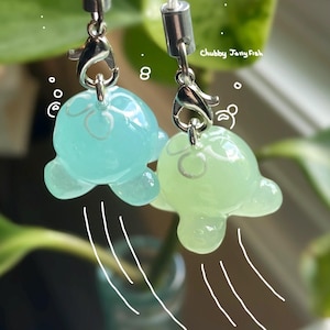 May include: Two translucent jellyfish charms in light blue and green, each suspended by a silver-tone clasp and white cord. The charms have a whimsical design, with the text "Chubby Jelly Fish" visible in the background.