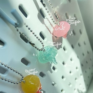 May include: Three jellyfish pendants on silver ball chains. The pendants are in pink, blue-green, and yellow, with the text "Pink Moon Jellyfish", "Moon Jellyfish", and "Blubber Jellyfish" written on the image.