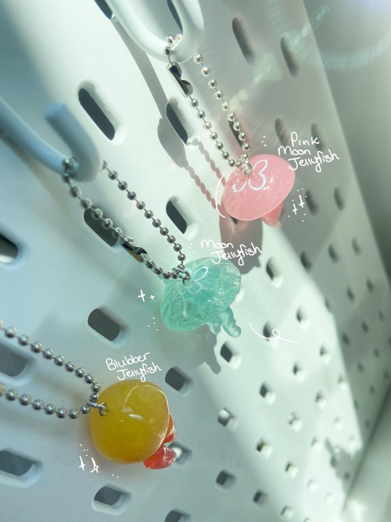 May include: Three jellyfish pendants on silver ball chains. The pendants are in pink, blue-green, and yellow, with the text "Pink Moon Jellyfish", "Moon Jellyfish", and "Blubber Jellyfish" written on the image.