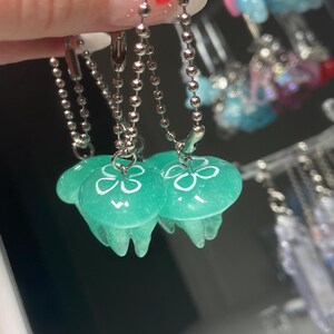 May include: Close-up of three translucent teal jellyfish charms with white floral designs, hanging from silver ball chains. The charms have a rounded top and flowing tentacles. Other colorful charms are visible in the background.
