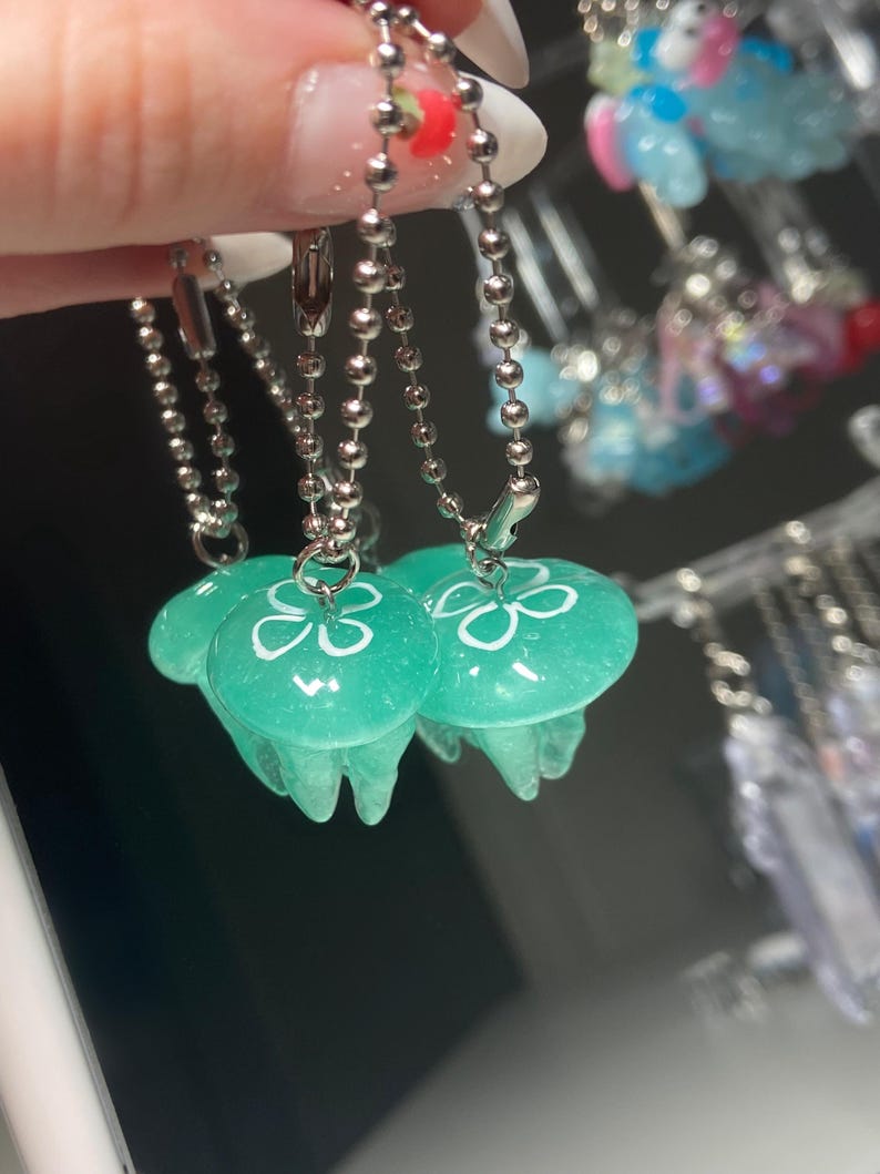 May include: Close-up of three translucent teal jellyfish charms with white floral designs, hanging from silver ball chains. The charms have a rounded top and flowing tentacles. Other colorful charms are visible in the background.