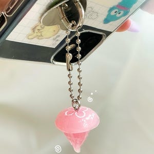 May include: A phone case with a silver ring holder and a pink jellyfish charm. The jellyfish charm is attached to the ring holder with a silver chain. The phone case has a cartoon design.