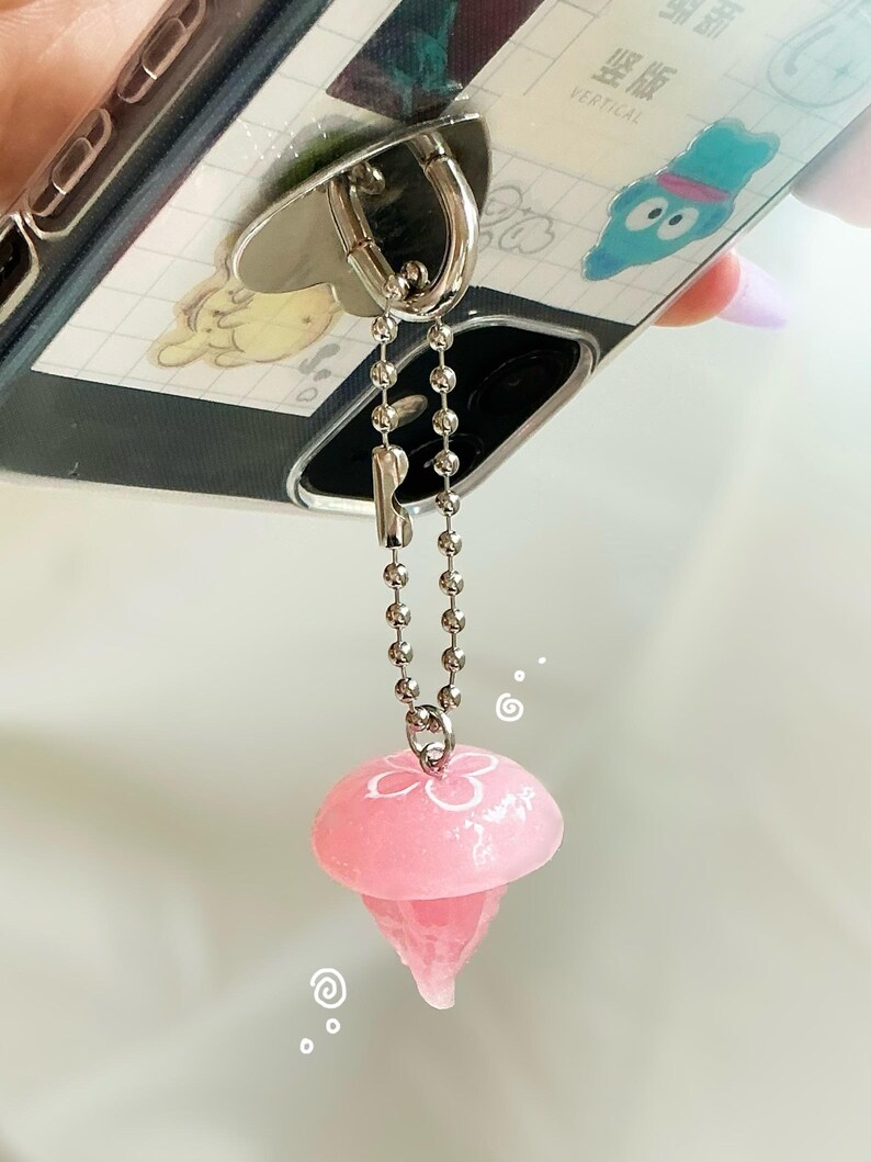 May include: A phone case with a silver ring holder and a pink jellyfish charm. The jellyfish charm is attached to the ring holder with a silver chain. The phone case has a cartoon design.