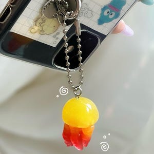 May include: A clear phone case with a silver heart-shaped ring holder and a decorative jellyfish charm. The charm is yellow, orange, and red, attached to a silver chain. The phone case has a white and gray design.