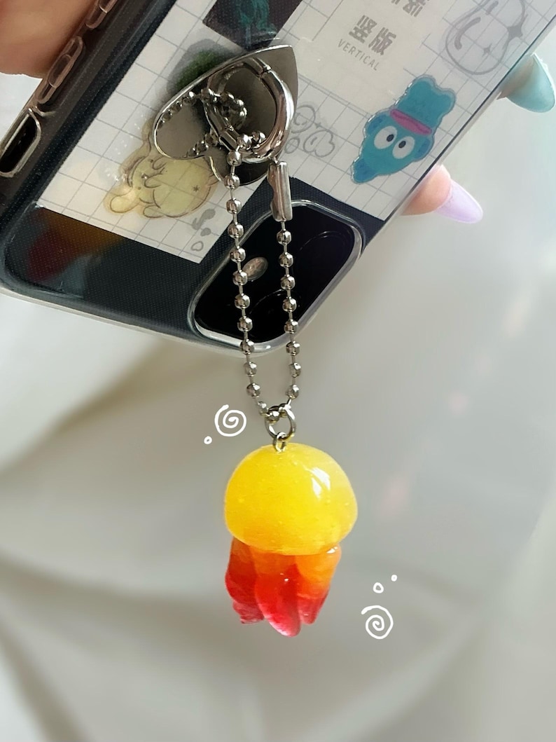 May include: A clear phone case with a silver heart-shaped ring holder and a decorative jellyfish charm. The charm is yellow, orange, and red, attached to a silver chain. The phone case has a white and gray design.