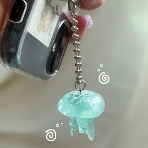 May include: A translucent, light blue jellyfish charm with a silver chain and clasp. The charm is attached to a clear phone case. The jellyfish has a rounded top and flowing tentacles. Decorative swirls are in the background.