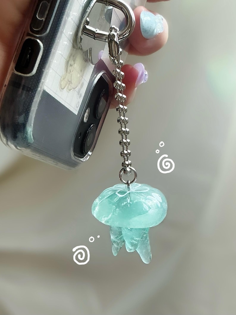 May include: A translucent, light blue jellyfish charm with a silver chain and clasp. The charm is attached to a clear phone case. The jellyfish has a rounded top and flowing tentacles. Decorative swirls are in the background.
