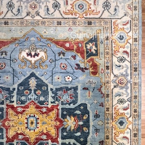 Puede incluir: Vista de esquina de una alfombra anudada a mano con un patrón geométrico detallado. La alfombra presenta un diseño central en tonos azules, rojos y dorados, con un borde crema. El texto "GulnajiHandKnots" es visible.