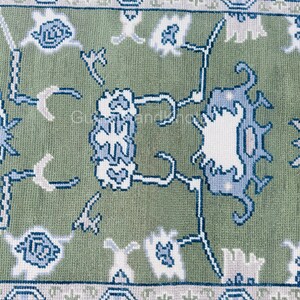 K&ouml;nnte beinhalten: Nahaufnahme eines gr&uuml;nen Teppichs mit detailliertem Blumenmuster in Wei&szlig; und Blau. Der Teppich hat einen Rand mit einem sich wiederholenden geometrischen Design. Der Text "GulnajHandKnots" ist in den Ecken sichtbar.
