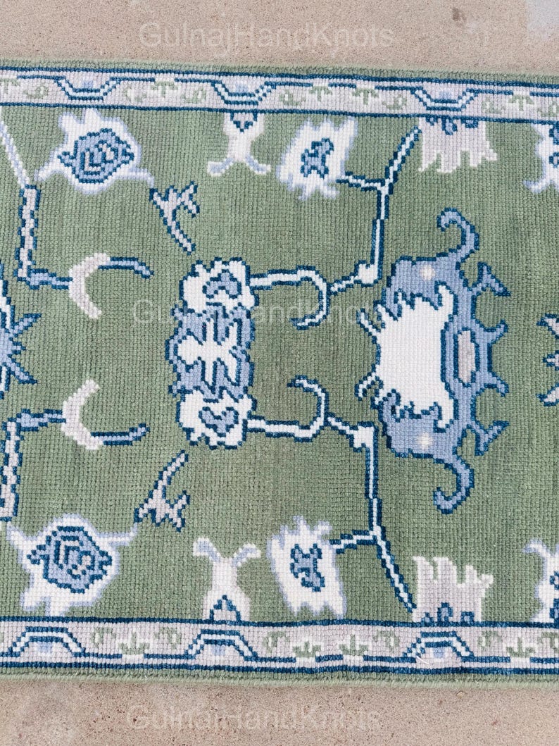 K&ouml;nnte beinhalten: Nahaufnahme eines gr&uuml;nen Teppichs mit detailliertem Blumenmuster in Wei&szlig; und Blau. Der Teppich hat einen Rand mit einem sich wiederholenden geometrischen Design. Der Text "GulnajHandKnots" ist in den Ecken sichtbar.