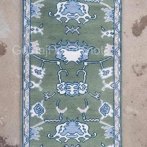 K&ouml;nnte beinhalten: Ein rechteckiger, gr&uuml;ner Teppich mit einem sich wiederholenden Muster aus blauen und wei&szlig;en floralen und geometrischen Designs. Der Teppich hat einen wei&szlig;en Fransen an den kurzen Enden und einen Rand mit einem &auml;hnlichen Design. Der Text "Gulnaj HandKnots" ist sichtbar.