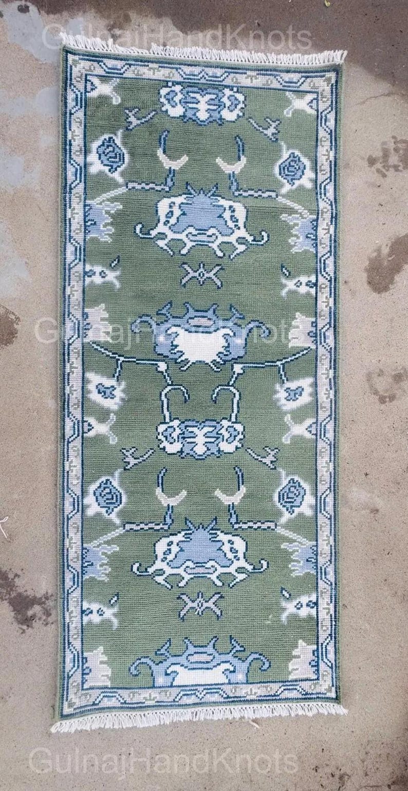 K&ouml;nnte beinhalten: Ein rechteckiger, gr&uuml;ner Teppich mit einem sich wiederholenden Muster aus blauen und wei&szlig;en floralen und geometrischen Designs. Der Teppich hat einen wei&szlig;en Fransen an den kurzen Enden und einen Rand mit einem &auml;hnlichen Design. Der Text "Gulnaj HandKnots" ist sichtbar.