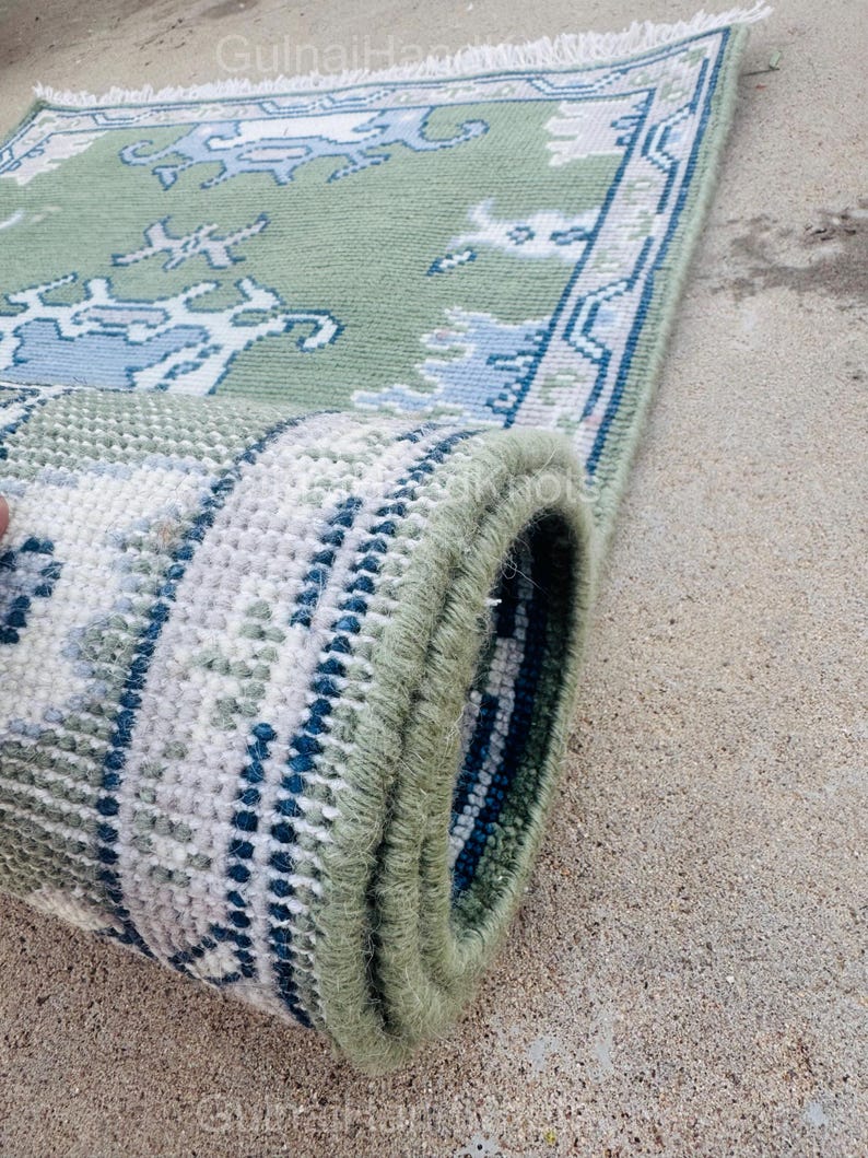 K&ouml;nnte beinhalten: Ein aufgerollter, rechteckiger Teppich mit gr&uuml;nem Hintergrund und einem sich wiederholenden Muster in Blau- und Wei&szlig;t&ouml;nen. Der Rand des Teppichs weist ein dunkleres blaues Design auf. Der Teppich hat an einem Ende Fransen.