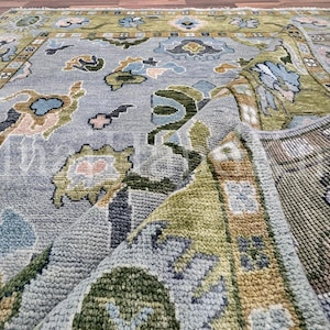 May include: A rectangular area rug featuring a light blue central field with a multi-colored floral and vine pattern in shades of olive green, tan, and dark blue. The rug has a wide olive green border and is folded to reveal the textured underside.