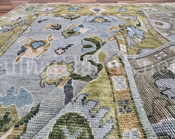 Könnte beinhalten: Ein rechteckiger Teppich mit einem hellblauen Mittelfeld und einem mehrfarbigen Blumen- und Rankenmuster in Olivgrün, Beige und Dunkelblau. Der Teppich hat einen breiten olivgrünen Rand und ist umgeschlagen, um die strukturierte Unterseite zu zeigen.