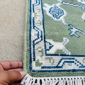 K&ouml;nnte beinhalten: Nahaufnahme eines gr&uuml;nen Teppichs mit detailliertem Rand in Blau, Wei&szlig; und Grau. Der Teppich hat ein Blumenmuster und einen Fransenrand. Der Teppich ist handgefertigt.