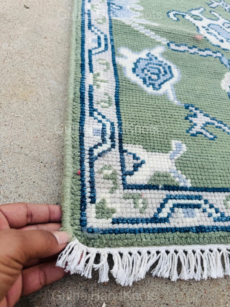 K&ouml;nnte beinhalten: Nahaufnahme eines gr&uuml;nen Teppichs mit detailliertem Rand in Blau, Wei&szlig; und Grau. Der Teppich hat ein Blumenmuster und einen Fransenrand. Der Teppich ist handgefertigt.