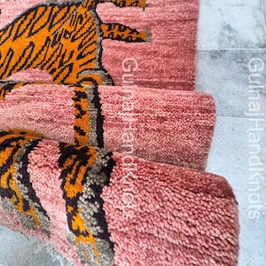 May include: A rolled-up, pink rug with a tiger design. The tiger is orange and black, with a detailed pattern. The rug has a textured, woven appearance, and the text "Gulnaj HandKnots" is visible on the side.